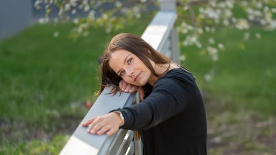 Naisoletettu tummahiuksinen nainen nojaa molemmilla käsillään kuvan vasemmalla puolella olevaan aitaan ja katsoo ilmeettömänä kameraan.